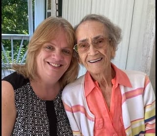 Two smiling women posing together indoors near a window with white frames and green foliage visible outside